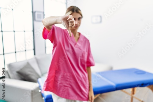 Young blonde woman working as physiotherapist at home looking unhappy and angry showing rejection and negative with thumbs down gesture. bad expression.