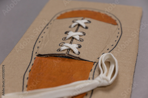 Card for tying laces is against gray background. White shoelace is inserted into the eyelets. The second shoelace lies next to it. Homemade card made of three-ply brown cardstock. Series part. photo