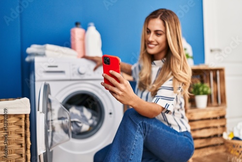 Young blonde woman using smartphone and washing clothes at laundry room