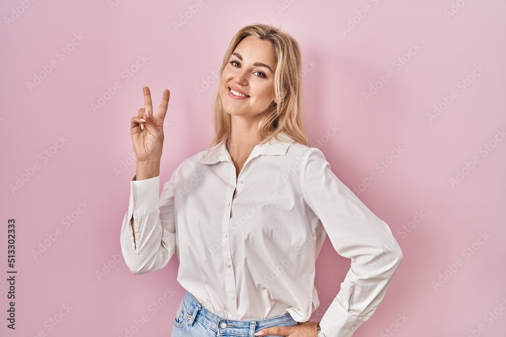 Young caucasian woman wearing casual white shirt over pink background smiling looking to the camera showing fingers doing victory sign. number two. Young caucasian woman wearing casual white shirt over pink background smiling looking to the camera showing fingers doing victory sign. number two.