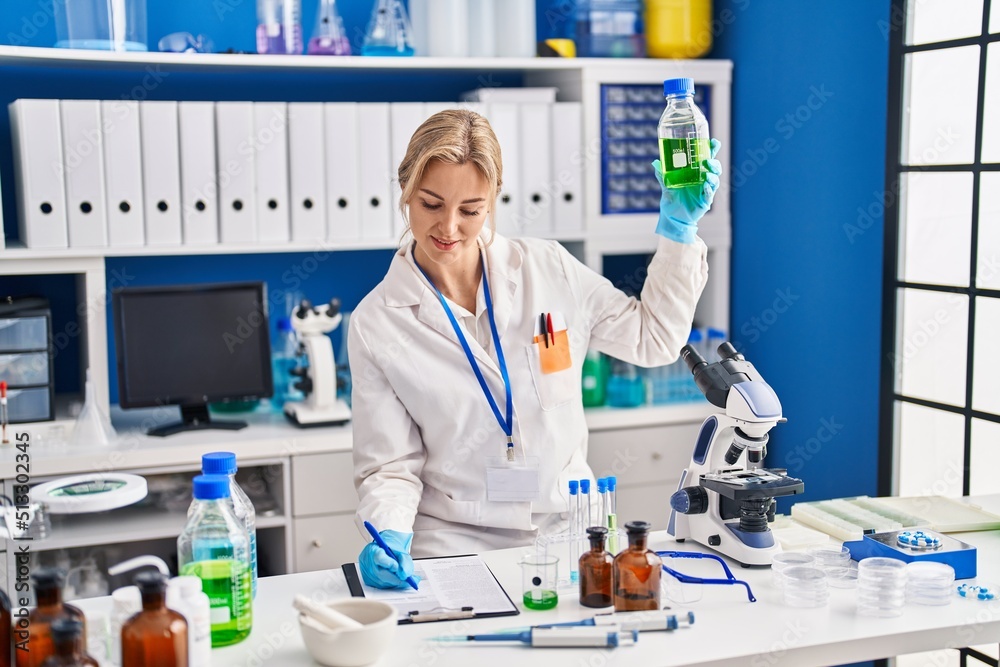 Young blonde woman scientist measuring liquid writing on document at laboratory
