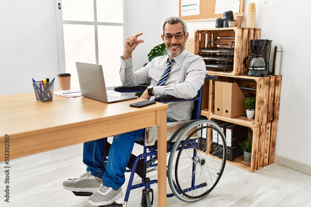 Middle age hispanic man working at the office sitting on wheelchair smiling and confident gesturing with hand doing small size sign with fingers looking and the camera. measure concept. Middle age hispanic man working at the office sitting on wheelchair smiling and confident gesturing with hand doing small size sign with fingers looking and the camera. measure concept.