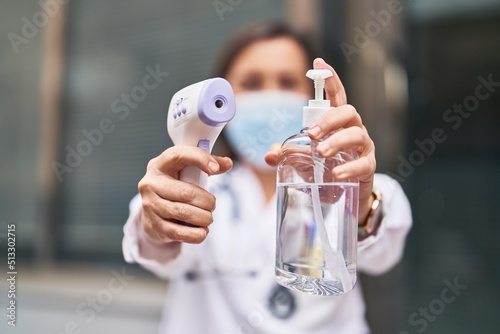Middle age woman wearing doctor uniform and medical mask holding thermometer and sanitizer gel hands at street
