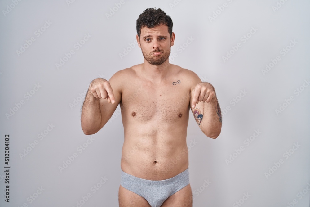 Young hispanic man standing shirtless wearing underware pointing down looking sad and upset, indicating direction with fingers, unhappy and depressed. Young hispanic man standing shirtless wearing underware pointing down looking sad and upset, indicating direction with fingers, unhappy and depressed.
