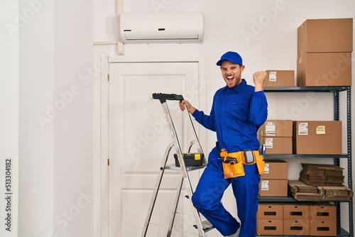 Young hispanic man working at renovation screaming proud, celebrating victory and success very excited with raised arms