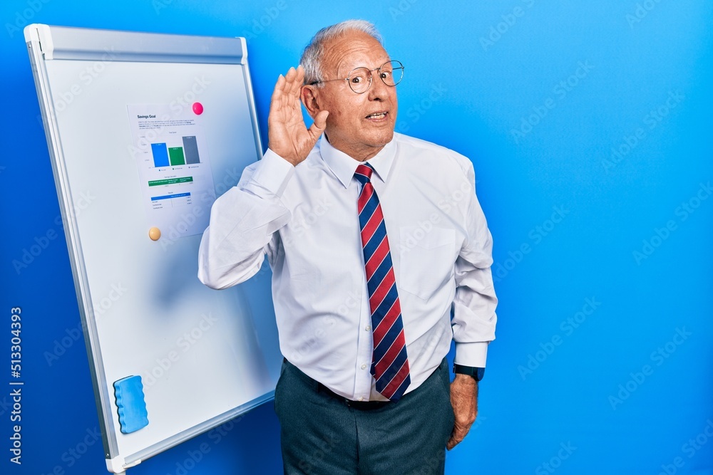 Senior man with grey hair standing by business blackboard smiling with hand over ear listening an hearing to rumor or gossip. deafness concept.