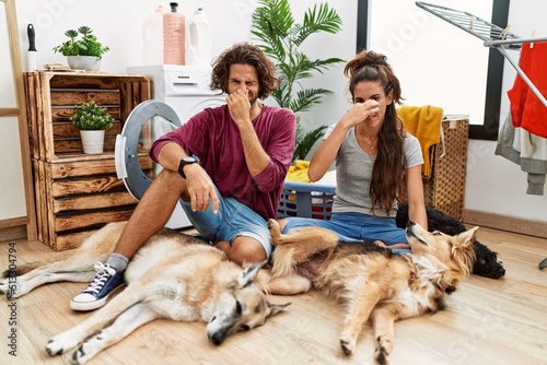 Young hispanic couple doing laundry with dogs smelling something stinky and disgusting, intolerable smell, holding breath with fingers on nose. bad smell