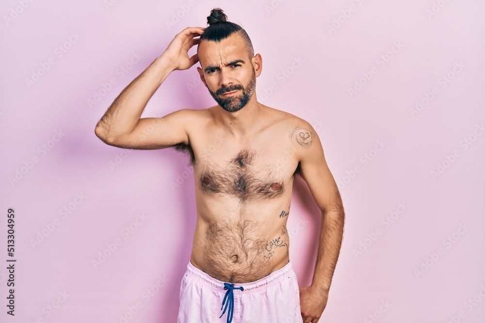 Fototapeta premium Young hispanic man shirtless wearing swimsuit confuse and wonder about question. uncertain with doubt, thinking with hand on head. pensive concept.