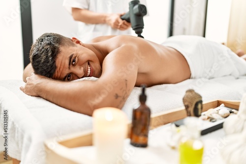 Young hispanic man relaxed having back massage using percussion pistol at beauty center