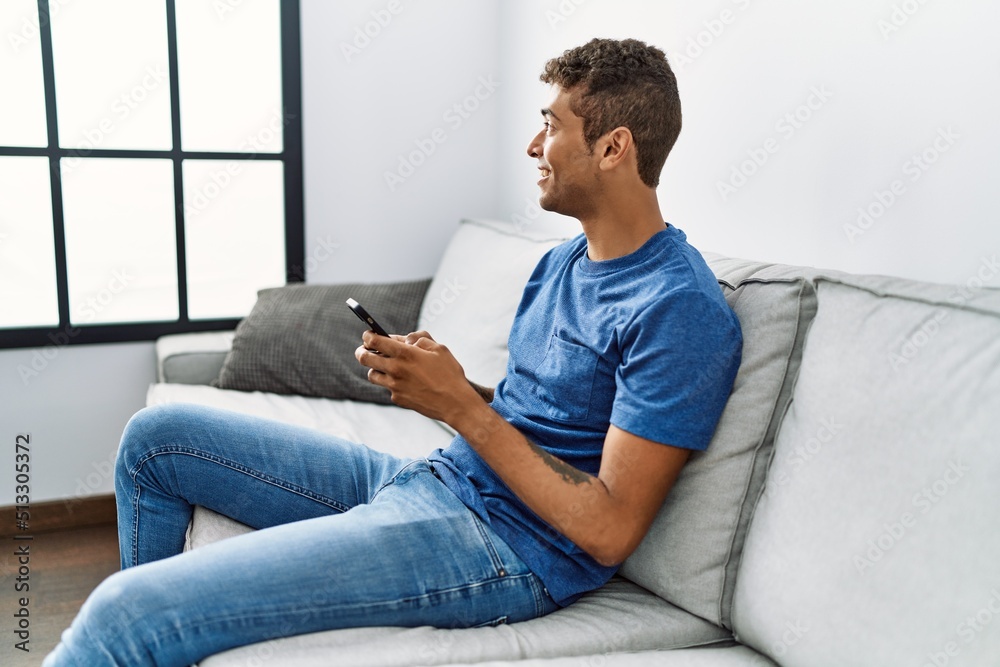 Young hispanic man relaxing sitting on the sofa using smartphone at home Young hispanic man relaxing sitting on the sofa using smartphone at home