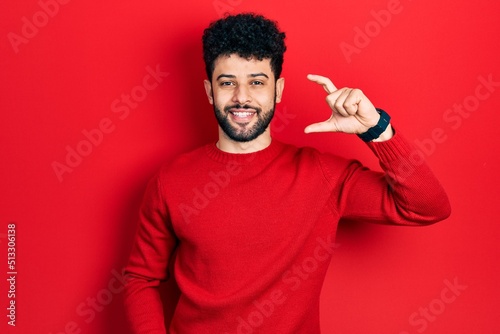 Young arab man with beard wearing casual red sweater smiling and confident gesturing with hand doing small size sign with fingers looking and the camera. measure concept.
