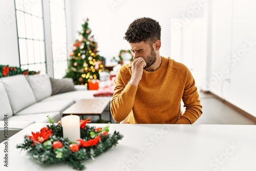 Arab young man sitting on the table by christmas tree smelling something stinky and disgusting  intolerable smell  holding breath with fingers on nose. bad smell