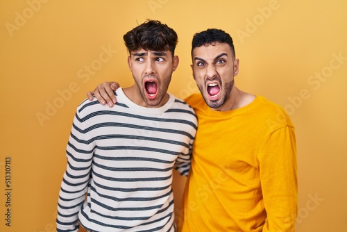 Young hispanic gay couple standing over yellow background angry and mad screaming frustrated and furious  shouting with anger. rage and aggressive concept.