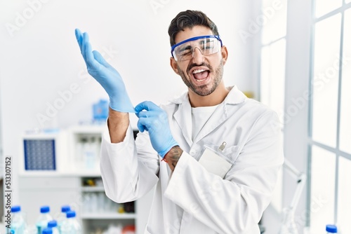 Young hispanic man working at scientist laboratory putting gloves on winking looking at the camera with sexy expression, cheerful and happy face.