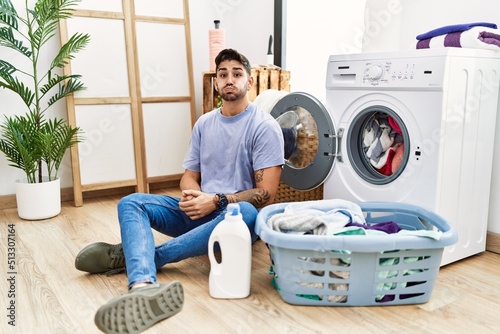 Young hispanic man putting dirty laundry into washing machine puffing cheeks with funny face. mouth inflated with air, crazy expression.