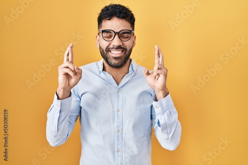 Hispanic man with beard standing over yellow background gesturing finger crossed smiling with hope and eyes closed. luck and superstitious concept.