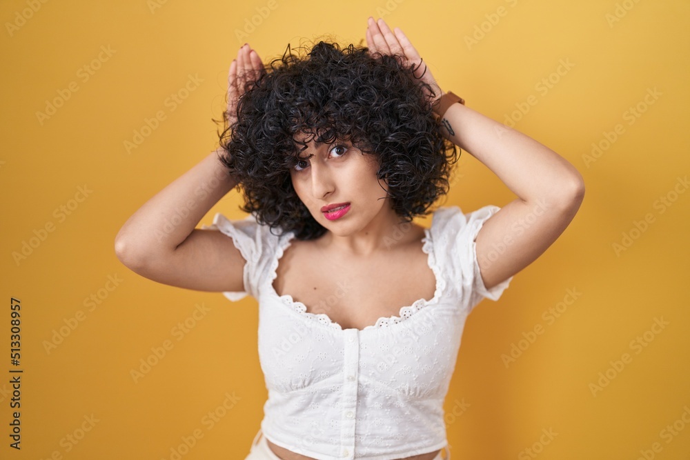Young brunette woman with curly hair standing over yellow background doing bunny ears gesture with hands palms looking cynical and skeptical. easter rabbit concept.