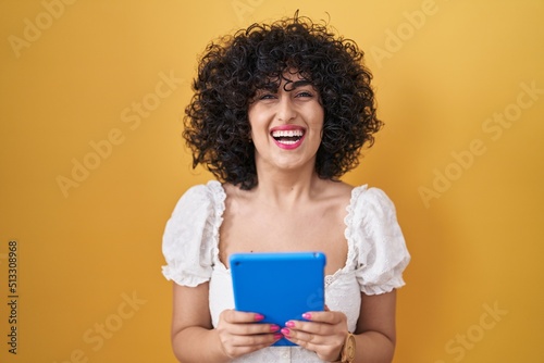 Young brunette woman with curly hair using touchpad over yellow background smiling and laughing hard out loud because funny crazy joke.