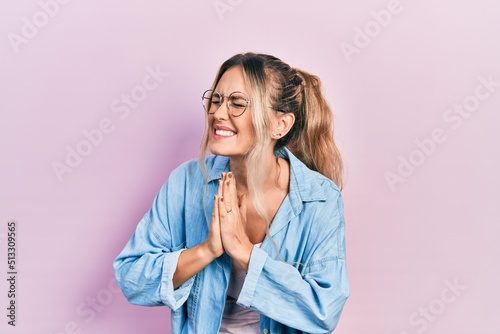 Beautiful young blonde woman wearing casual clothes and glasses begging and praying with hands together with hope expression on face very emotional and worried. begging. photo