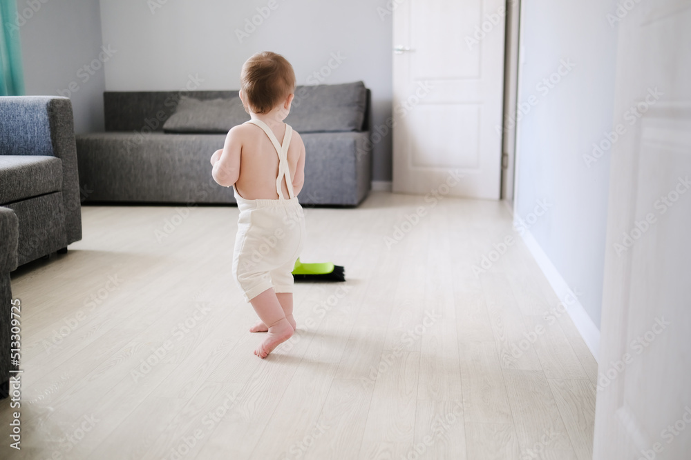 Toddler 1 year old with broom sweeps floor in bright room in Scandinavian style, montessori development, helping his mother and independence