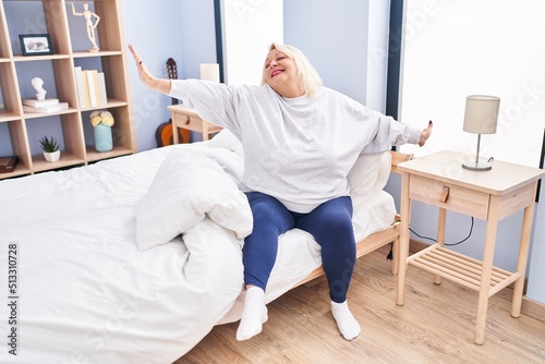Middle age blonde woman waking up stretching arms at bedroom
