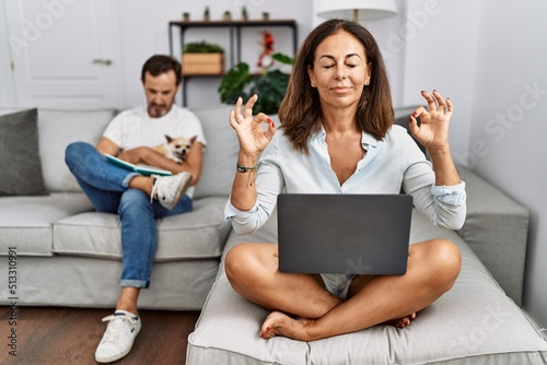 Hispanic middle age couple at home, woman using laptop relax and smiling with eyes closed doing meditation gesture with fingers. yoga concept.