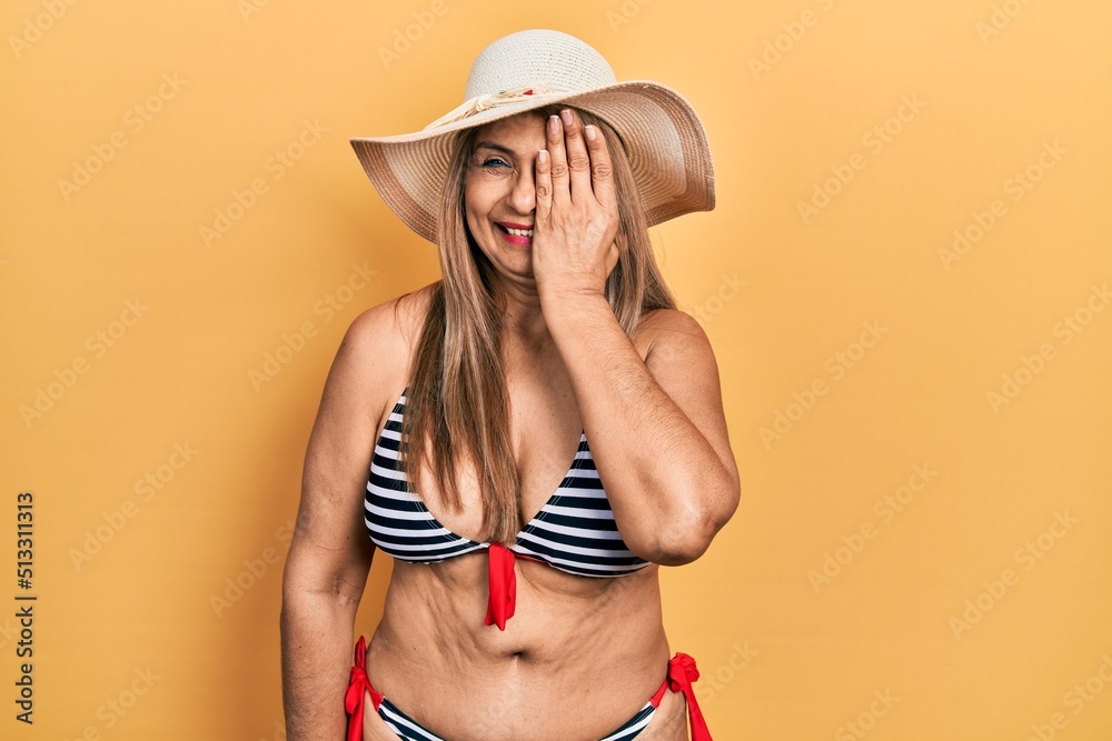 Middle age hispanic woman wearing bikini and summer hat covering one eye with hand, confident smile on face and surprise emotion. Middle age hispanic woman wearing bikini and summer hat covering one eye with hand, confident smile on face and surprise emotion.