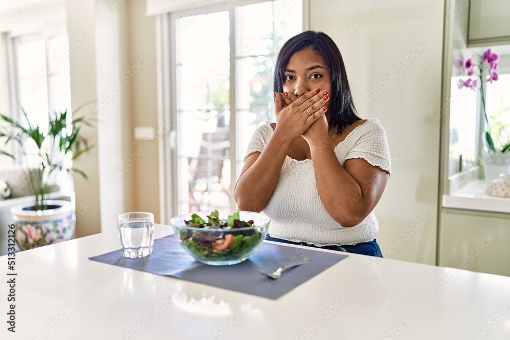 Young hispanic woman eating healthy salad at home shocked covering mouth with hands for mistake. secret concept.