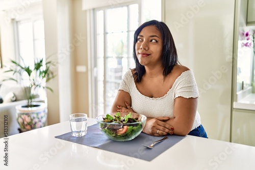 Young hispanic woman eating healthy salad at home smiling looking to the side and staring away thinking.