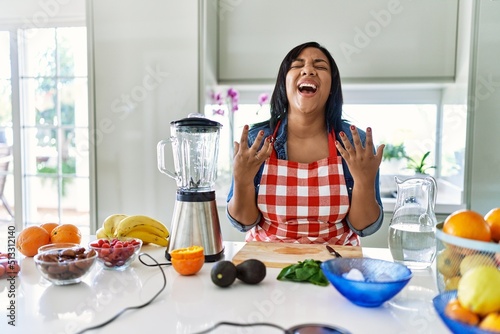 Young hispanic woman making healthy smoothie crazy and mad shouting and yelling with aggressive expression and arms raised. frustration concept.