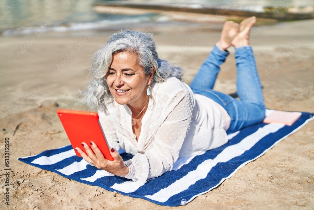 Middle age woman using touchpad lying on towel at seaside