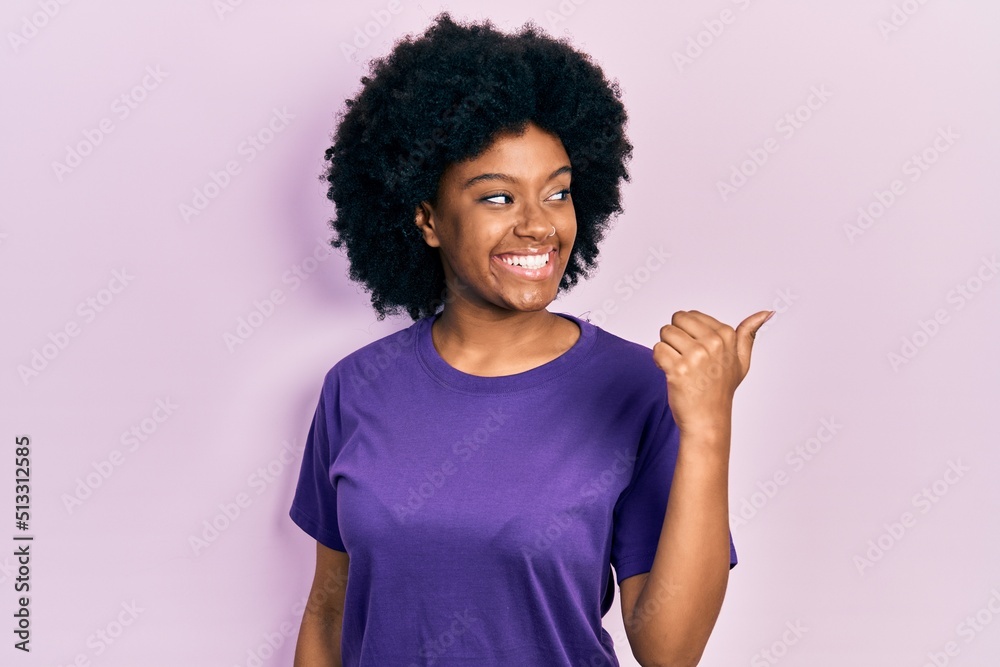 Young african american woman wearing casual clothes smiling with happy face looking and pointing to the side with thumb up. Young african american woman wearing casual clothes smiling with happy face looking and pointing to the side with thumb up.