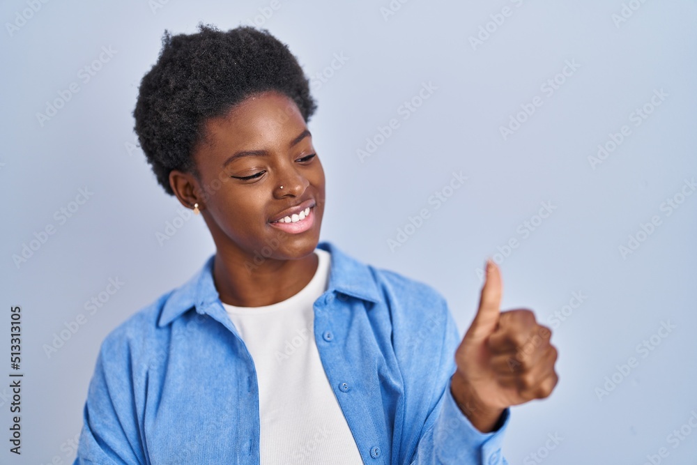 African american woman standing over blue background looking proud, smiling doing thumbs up gesture to the side African american woman standing over blue background looking proud, smiling doing thumbs up gesture to the side