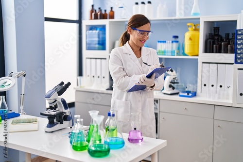 Middle age woman wearing scientist uniform writing on clipboard at laboratory