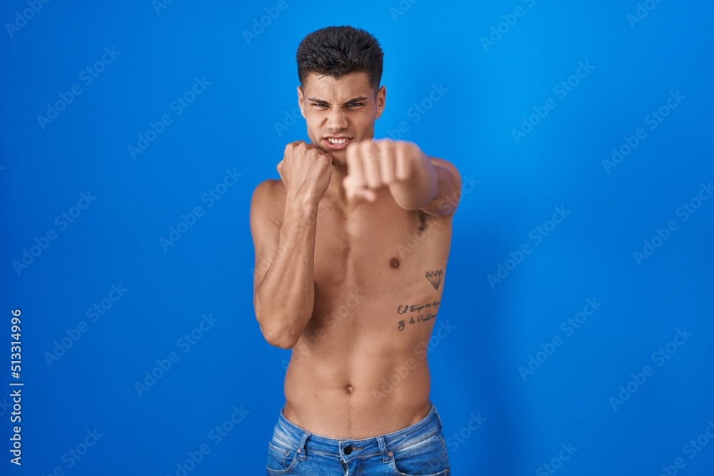 Fototapeta premium Young hispanic man standing shirtless over blue background punching fist to fight, aggressive and angry attack, threat and violence
