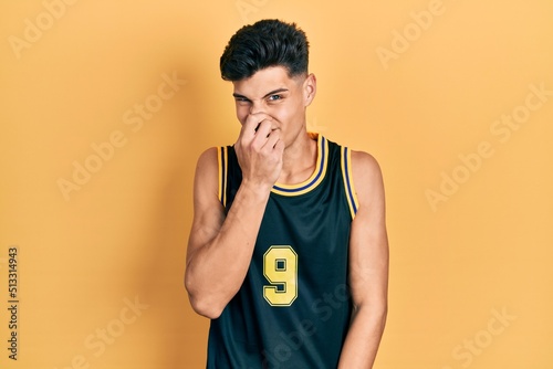 Young hispanic man wearing basketball uniform smelling something stinky and disgusting, intolerable smell, holding breath with fingers on nose. bad smell
