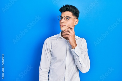 Young hispanic man wearing casual clothes and glasses serious face thinking about question with hand on chin, thoughtful about confusing idea