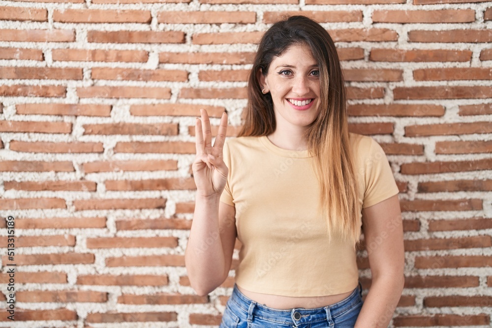 Young brunette woman standing over bricks wall showing and pointing up with fingers number three while smiling confident and happy. Young brunette woman standing over bricks wall showing and pointing up with fingers number three while smiling confident and happy.