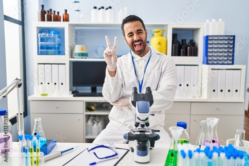 Young hispanic man with beard working at scientist laboratory smiling with happy face winking at the camera doing victory sign. number two.