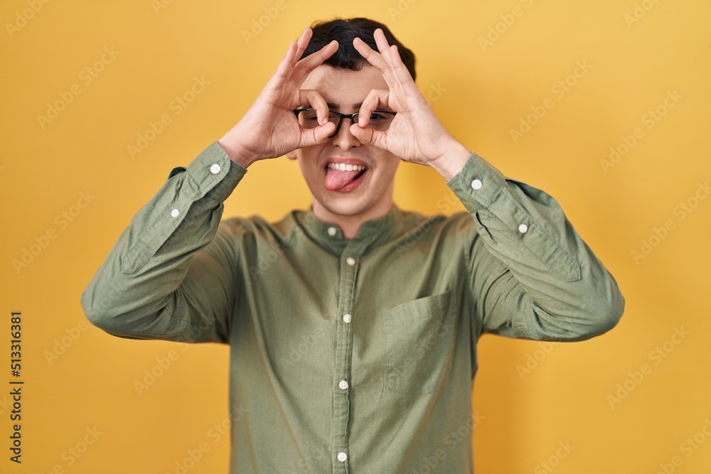 Non binary person standing over yellow background doing ok gesture like binoculars sticking tongue out, eyes looking through fingers. crazy expression. Non binary person standing over yellow background doing ok gesture like binoculars sticking tongue out, eyes looking through fingers. crazy expression.