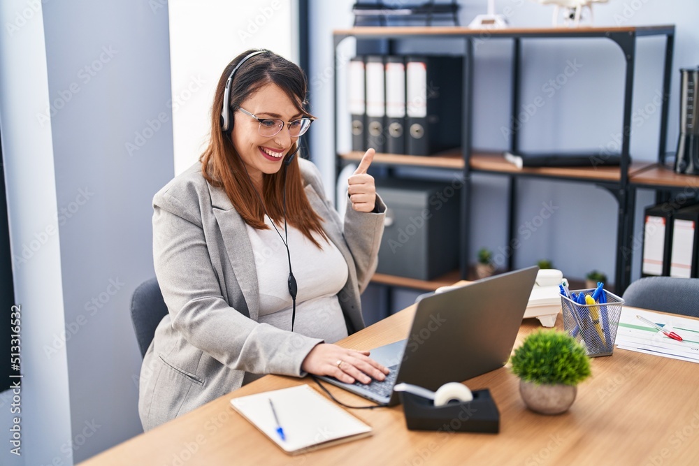 Pregnant woman working at the office wearing operator headset smiling happy and positive, thumb up doing excellent and approval sign Pregnant woman working at the office wearing operator headset smiling happy and positive, thumb up doing excellent and approval sign