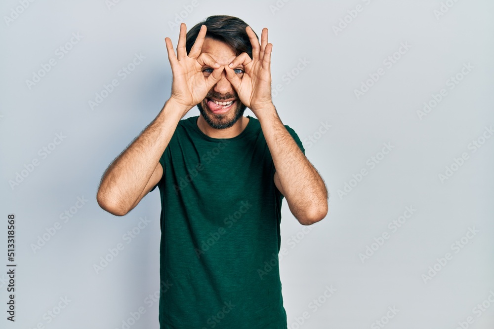 Young hispanic man wearing casual white tshirt doing ok gesture like binoculars sticking tongue out, eyes looking through fingers. crazy expression. Young hispanic man wearing casual white tshirt doing ok gesture like binoculars sticking tongue out, eyes looking through fingers. crazy expression.