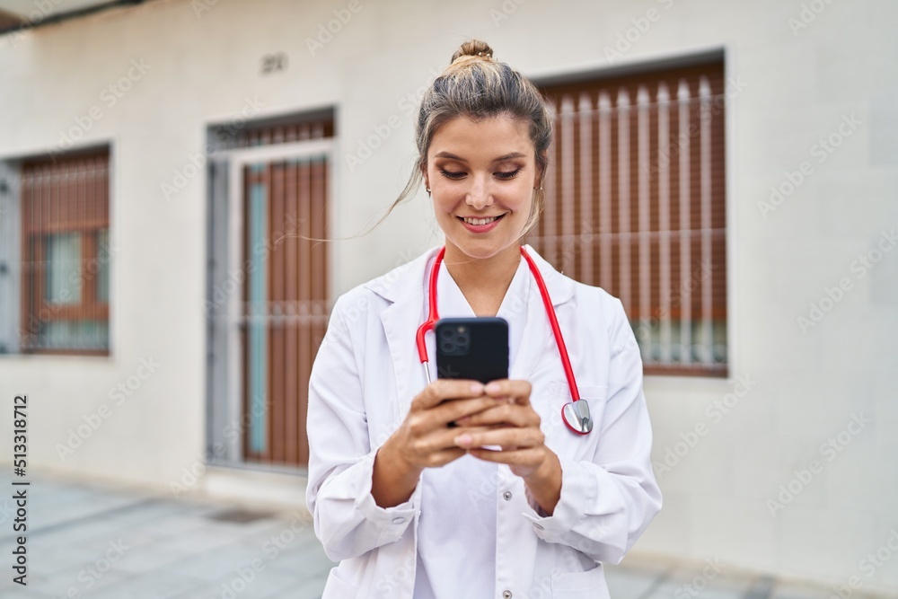 Fototapeta premium Young blonde woman wearing doctor uniform using smartphone at street