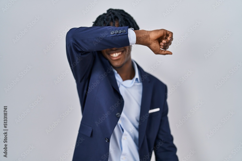 Young african man with dreadlocks wearing business jacket over white background covering eyes with arm smiling cheerful and funny. blind concept.