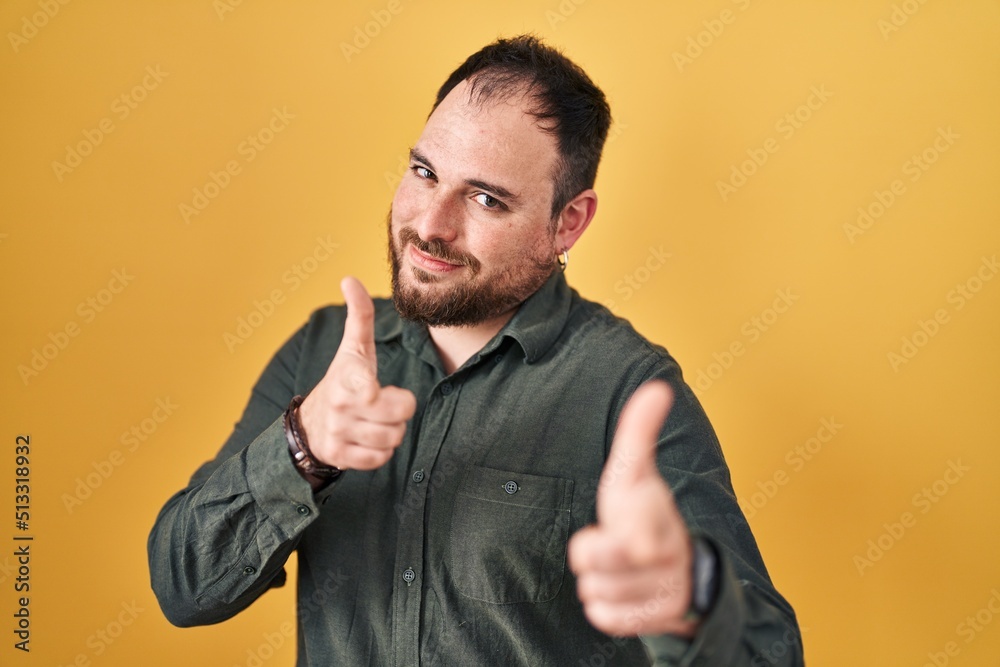 Plus size hispanic man with beard standing over yellow background pointing fingers to camera with happy and funny face. good energy and vibes. Plus size hispanic man with beard standing over yellow background pointing fingers to camera with happy and funny face. good energy and vibes.