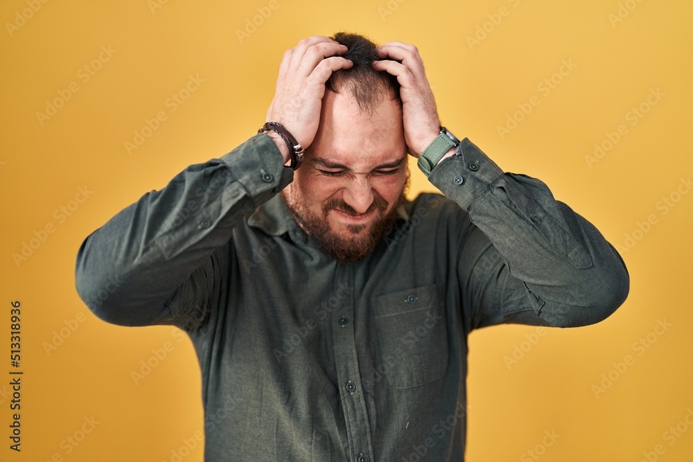 Plus size hispanic man with beard standing over yellow background suffering from headache desperate and stressed because pain and migraine. hands on head.