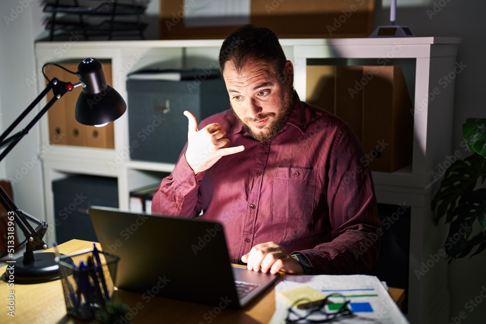 Plus size hispanic man with beard working at the office at night smiling doing phone gesture with hand and fingers like talking on the telephone. communicating concepts. Plus size hispanic man with beard working at the office at night smiling doing phone gesture with hand and fingers like talking on the telephone. communicating concepts.