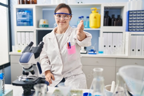 Hispanic girl with down syndrome working at scientist laboratory smiling cheerful offering palm hand giving assistance and acceptance.