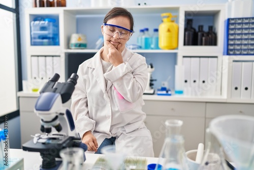 Hispanic girl with down syndrome working at scientist laboratory looking confident at the camera smiling with crossed arms and hand raised on chin. thinking positive.