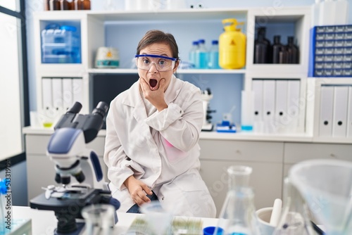 Hispanic girl with down syndrome working at scientist laboratory looking fascinated with disbelief  surprise and amazed expression with hands on chin